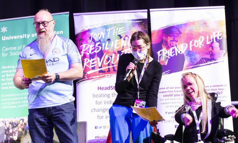 Three people are standing in front of in front of brightly coloured banners on a stage. The person in the middle is wearing a mask and singing into a microphone, on the right is a person in a wheelchair and the person on the left is wearing glasses and has a beard.