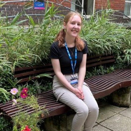 Photo of a woman sitting on a bench in a garden, smiling at the camera. She is wearing a black top and beige trousers, and has blonde hair.